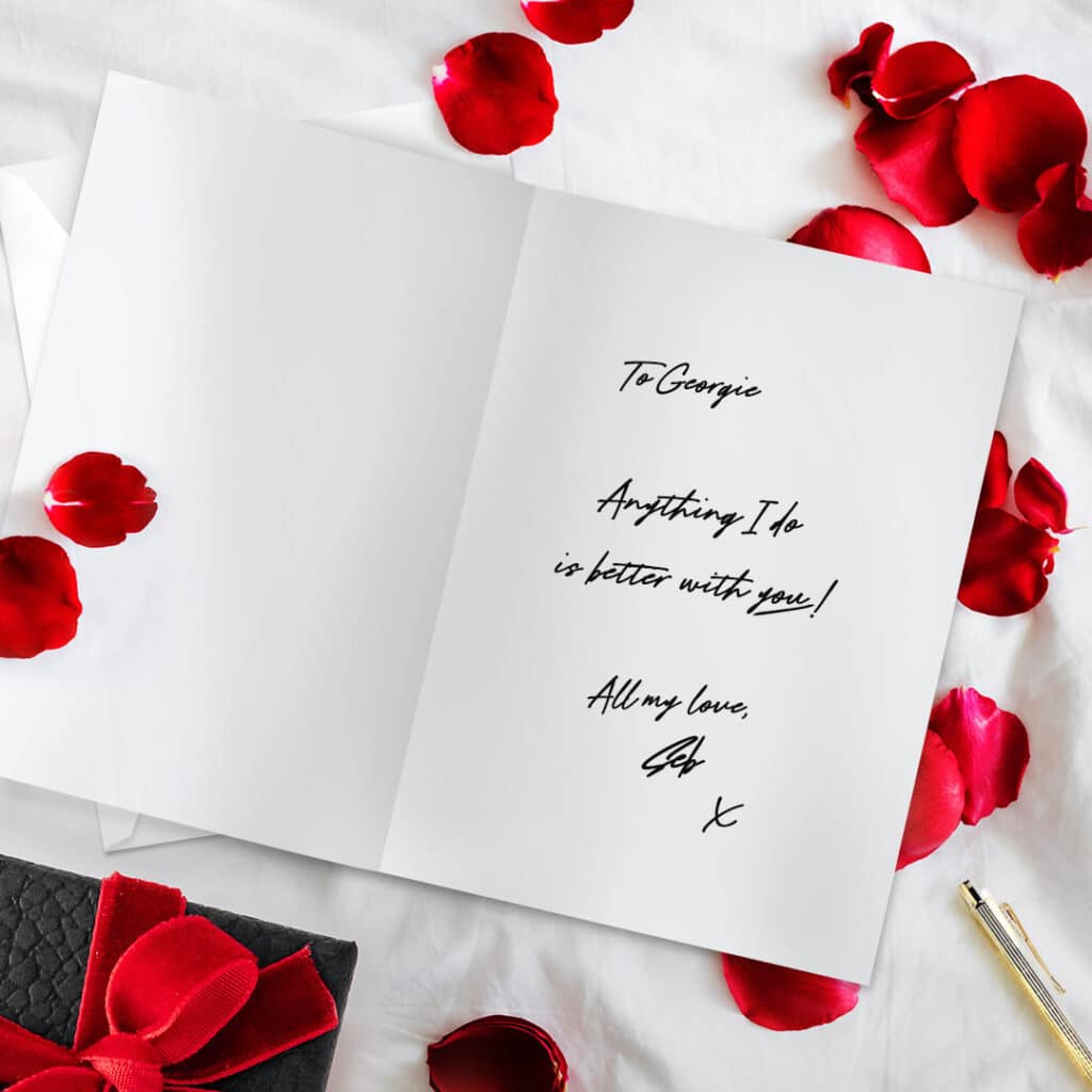 Open card with a love message inside on top of rose petals and beside a gift