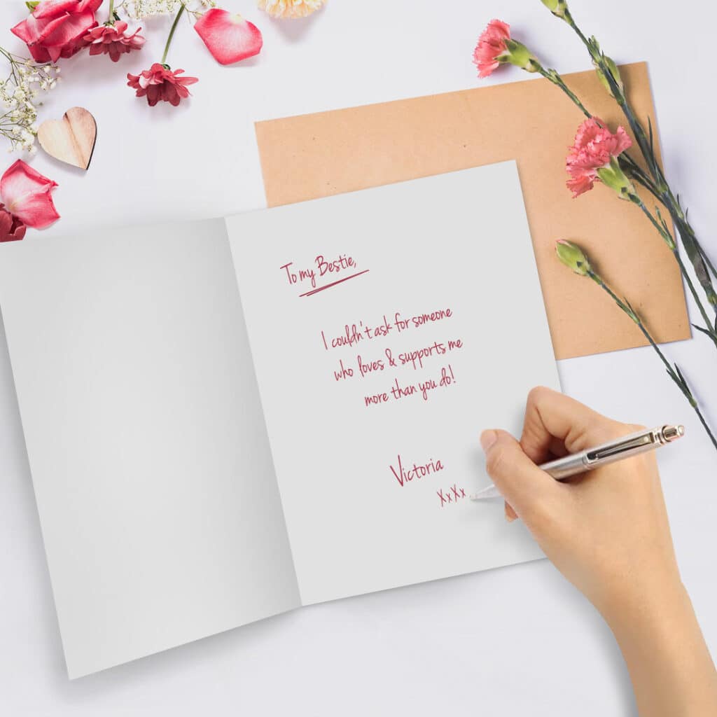 Open card beside flowers with a hand writing a love message inside with a pen